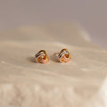 Trinity Knot Studs with an intertwined design in gold, silver, and rose gold tones rest on a beige surface—an ideal choice for a thoughtful anniversary gift.