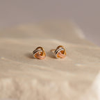 Trinity Knot Studs with an intertwined design in gold, silver, and rose gold tones rest on a beige surface—an ideal choice for a thoughtful anniversary gift.