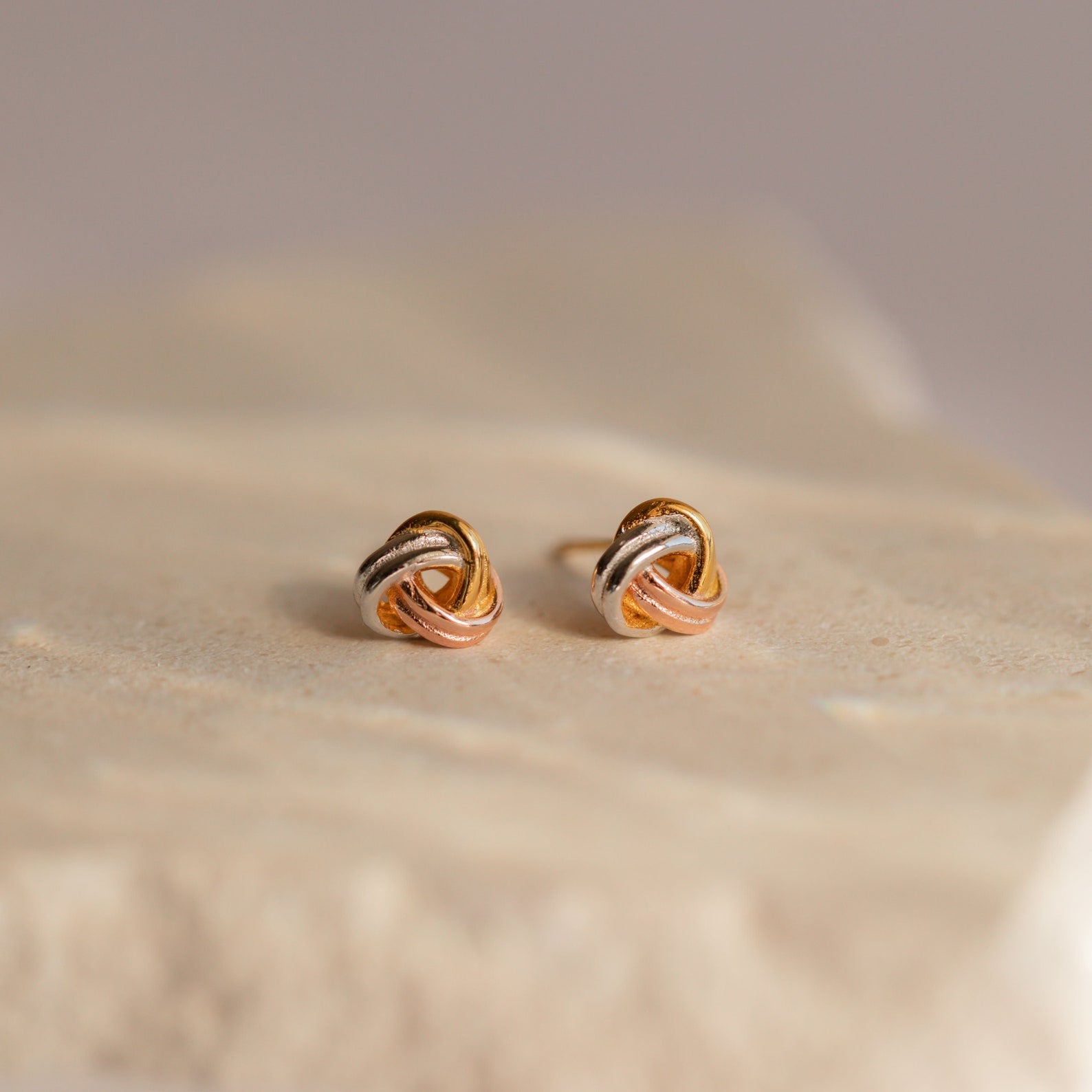 Trinity Knot Studs with an intertwined design in gold, silver, and rose gold tones rest on a beige surface—an ideal choice for a thoughtful anniversary gift.