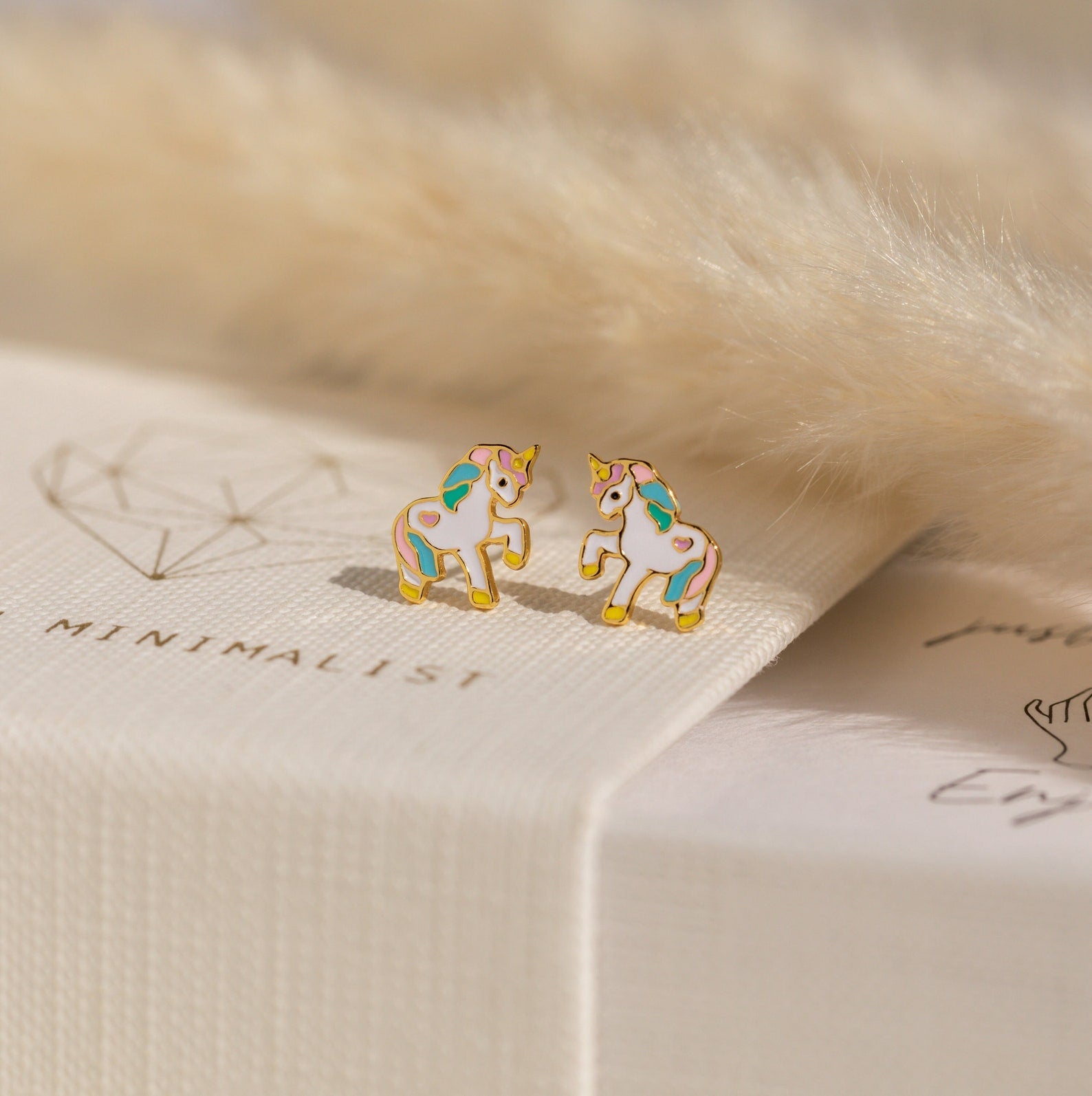 Unicorn Studs—small, colorful earrings—are displayed on a textured white surface next to soft beige faux fur, making them a charming gift for girls.