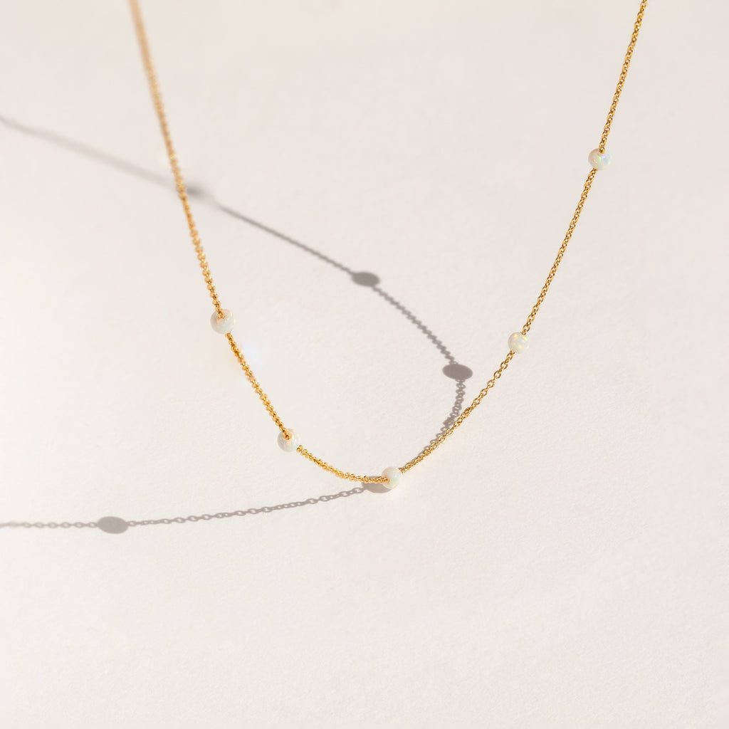Delicate gold chain necklace with evenly spaced small opal beads, shown casting soft shadows on a light cream background.
