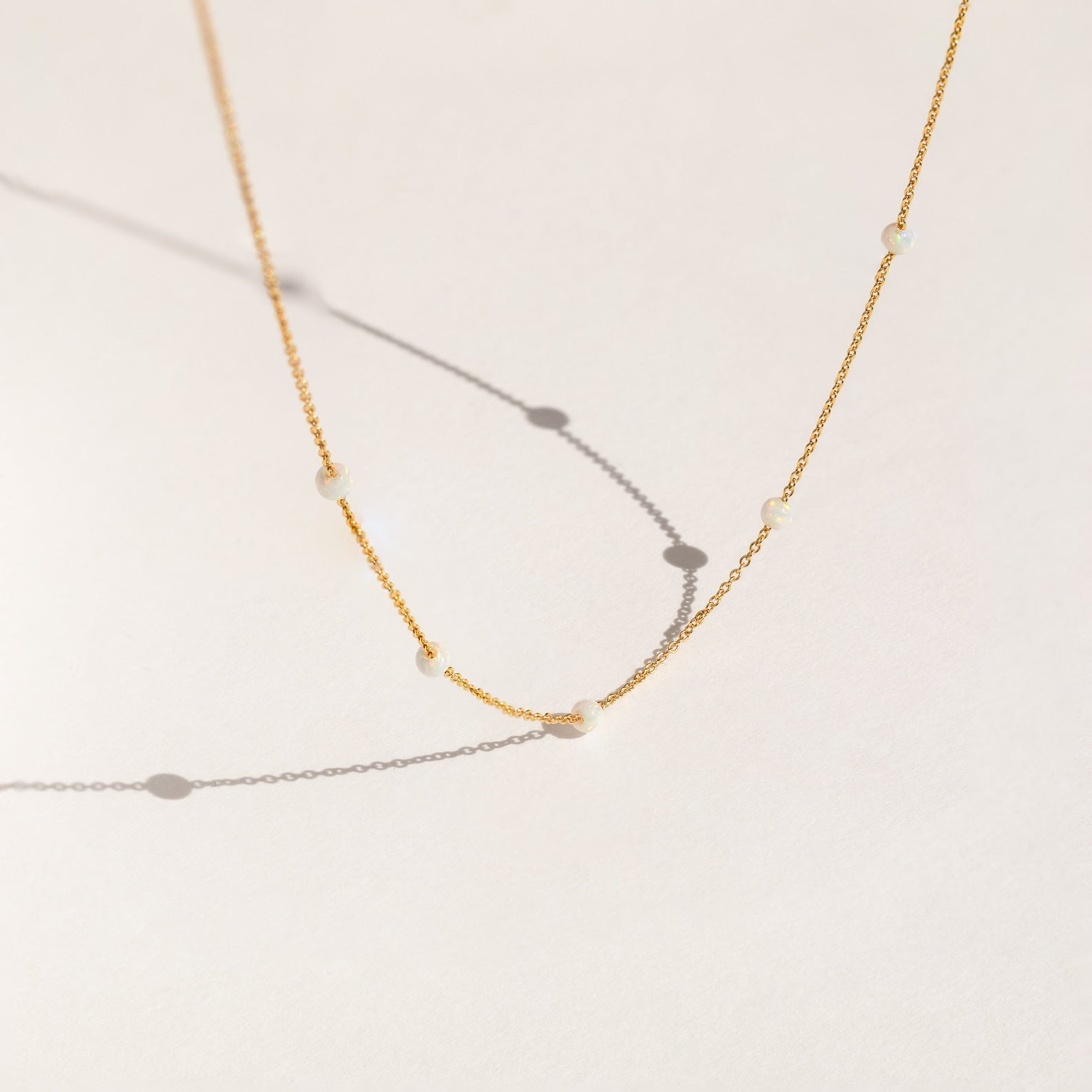 Delicate gold chain necklace with evenly spaced small opal beads, shown casting soft shadows on a light cream background.