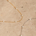 The Opal Station Necklace features gold and silver chains with small white opals in a minimalist design, showcased on a beige textured surface.