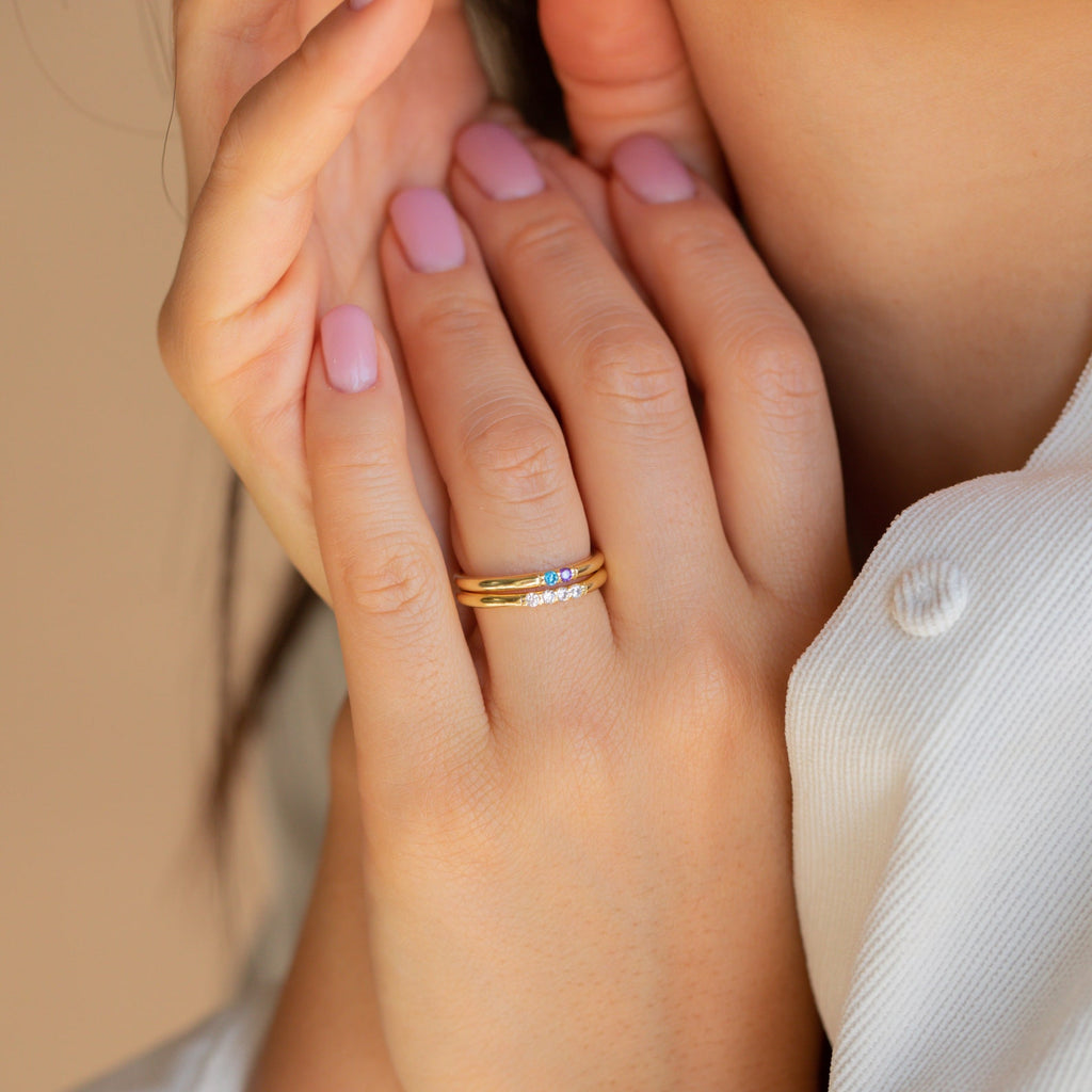 A hand with pink nails wears the Mini Birthstone Ring in Rose Gold along with a gold band featuring small diamonds, both near the face and neck.