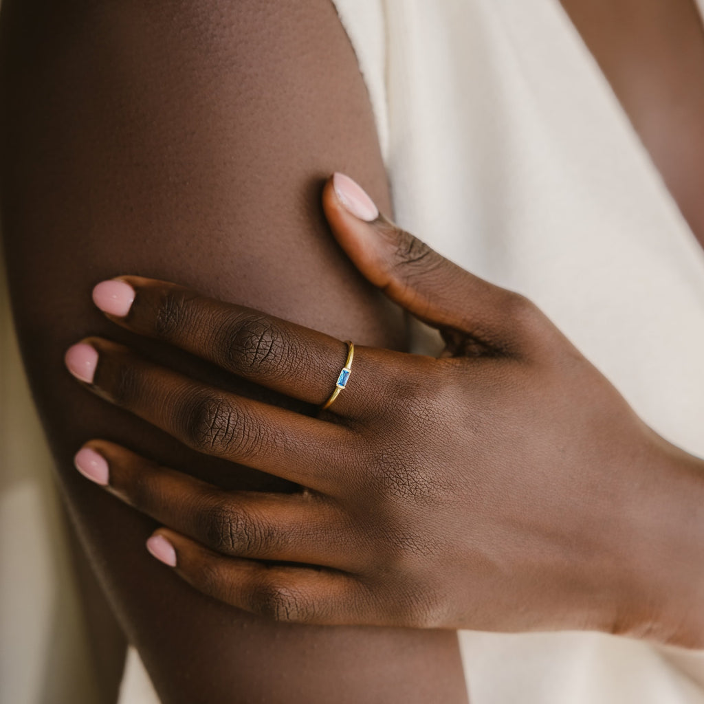 A hand with pale pink nails wears a Baguette Birthstone Ring with a blue stone, resting on a bare arm in soft light.