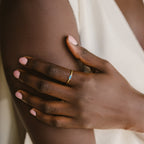 A hand with pale pink nails wears a Baguette Birthstone Ring with a blue stone, resting on a bare arm in soft light.