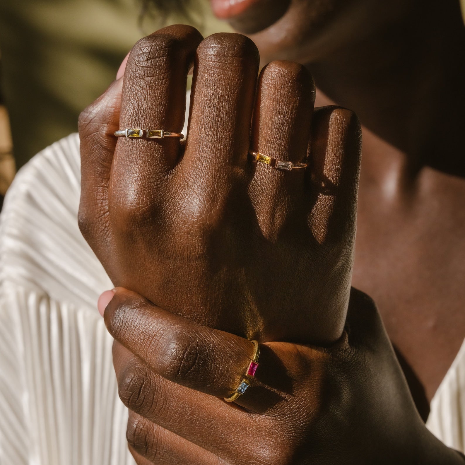 Close-up of hands with dark skin wearing gold rings, including the Duo Baguette Birthstone Ring with customized gemstones, against a light background—an ideal personalized gift.