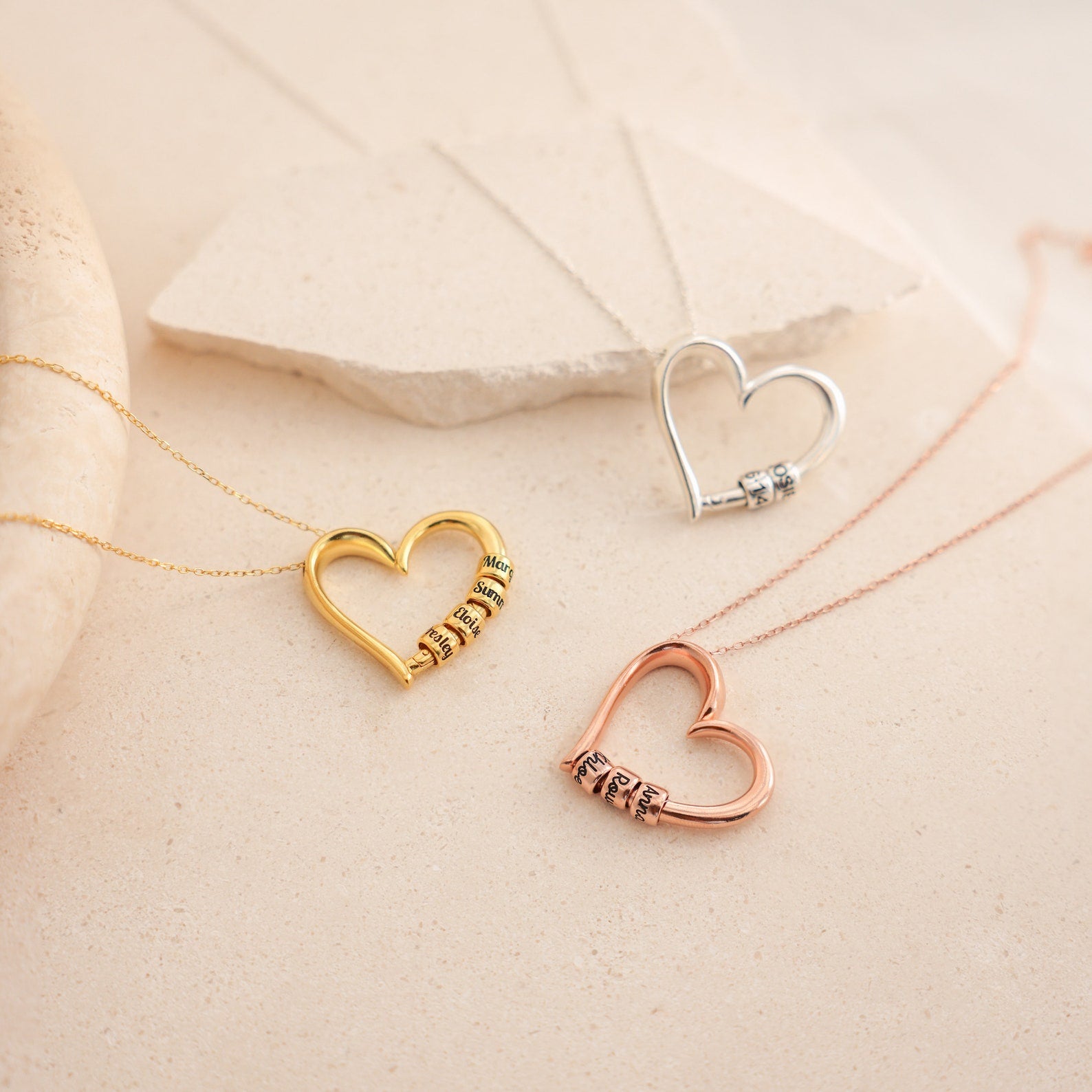 The Custom Bead Heart Necklace is shown in gold, silver, and rose gold. Each heart-shaped pendant features personalized engraved beads and is displayed on a beige surface.