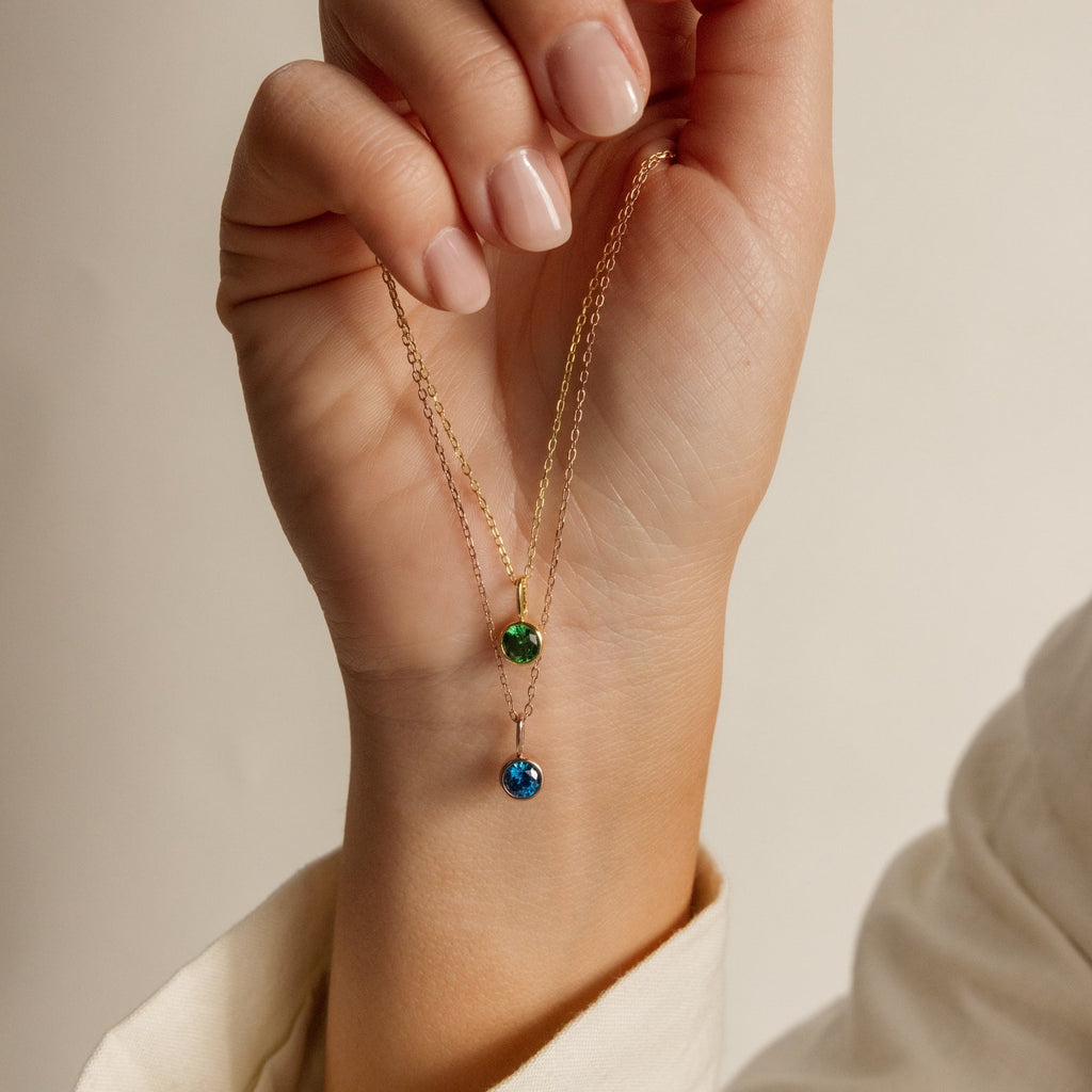 A hand holds two dainty Custom Birthstone Necklaces, each featuring a small green or blue gemstone pendant—perfect for a meaningful and personalized gift.