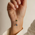 A hand holds two dainty Custom Birthstone Necklaces, each featuring a small green or blue gemstone pendant—perfect for a meaningful and personalized gift.
