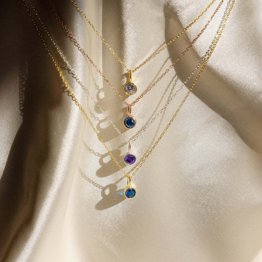 Five delicate gold Custom Birthstone Necklaces with colorful gemstone pendants—including a dainty necklace and birthstone styles—displayed on soft, cream-colored fabric.