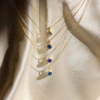 Five delicate gold Custom Birthstone Necklaces with colorful gemstone pendants—including a dainty necklace and birthstone styles—displayed on soft, cream-colored fabric.