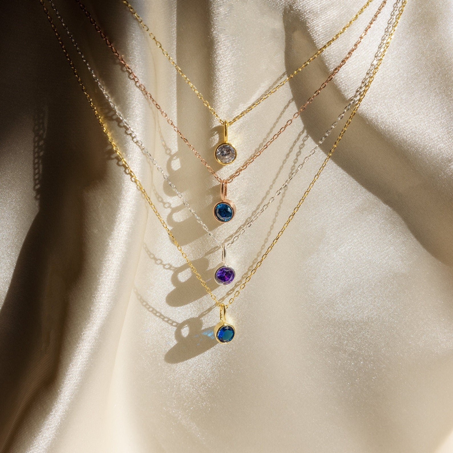 Five delicate gold Custom Birthstone Necklaces with colorful gemstone pendants—including a dainty necklace and birthstone styles—displayed on soft, cream-colored fabric.