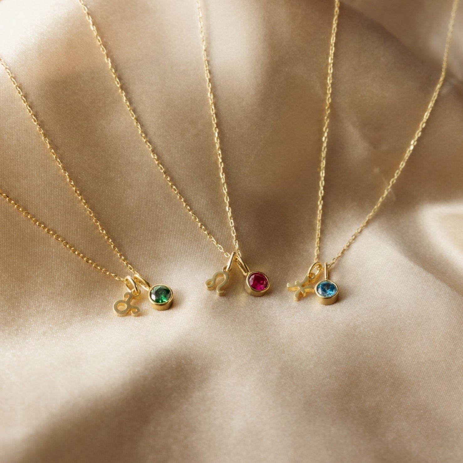 Four Zodiac Birthstone Necklaces with vibrant gemstone pendants and zodiac charms rest on beige satin—an ideal personalized jewelry gift.