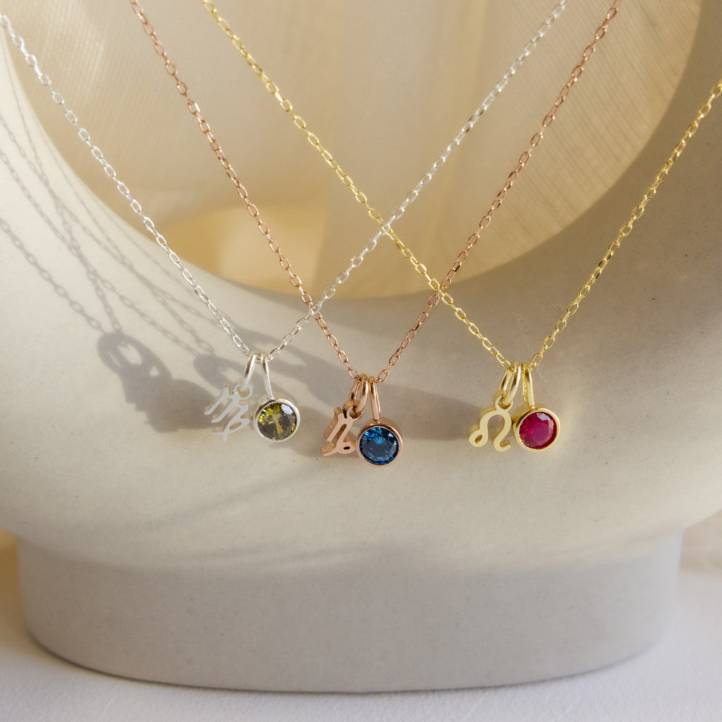 The Zodiac Birthstone Necklace features colored gemstones on silver, gold, and rose gold chains, elegantly displayed on a white curved surface—an ideal personalized jewelry or birthstone necklace gift.