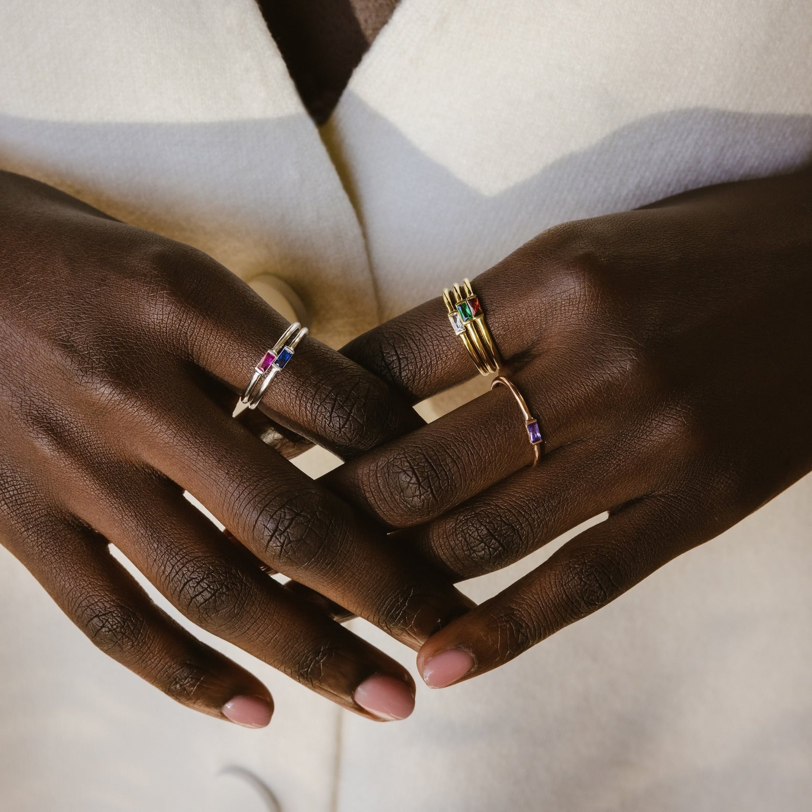 Close-up of hands wearing the Baguette Birthstone Ring alongside vibrant gemstone stacking rings, elegantly paired with a cream-colored coat.