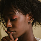A woman with closed eyes, wearing the Editor Huggie Earrings Set and pink eyeshadow, gently touches her lips.