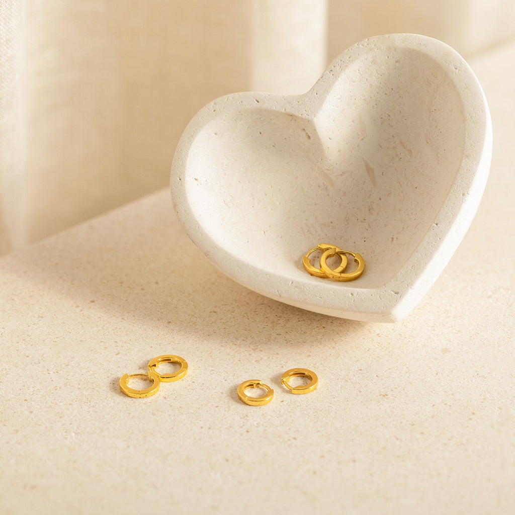 The Editor Huggie Earrings Set—small, dainty hoops displayed in and beside a heart-shaped, cream-colored stone dish on a beige surface—offers perfect minimalist style for everyday elegance.