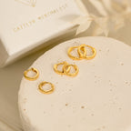 The Editor Huggie Earrings Set, featuring four pairs of minimalist hoops, is displayed on a textured white surface near a jewelry box—perfect for lovers of dainty earrings.