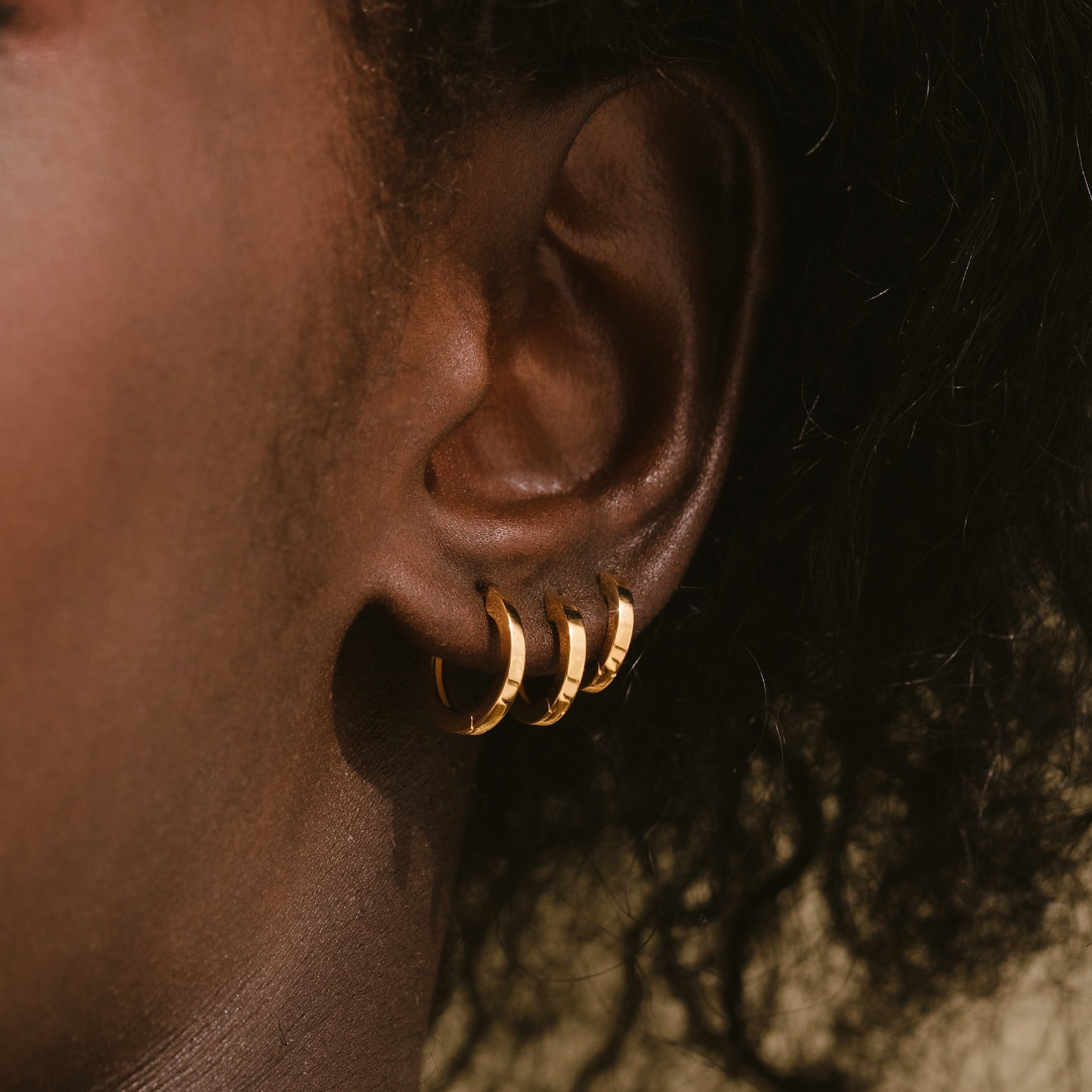 Close-up of a person’s ear wearing the Editor Huggie Earrings Set—three dainty hoops in the earlobe, shown against a neutral background.