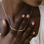 A hand with pink nails touches a Morse Code Necklace with silver beads on someone’s neck, paired with a white textured top.