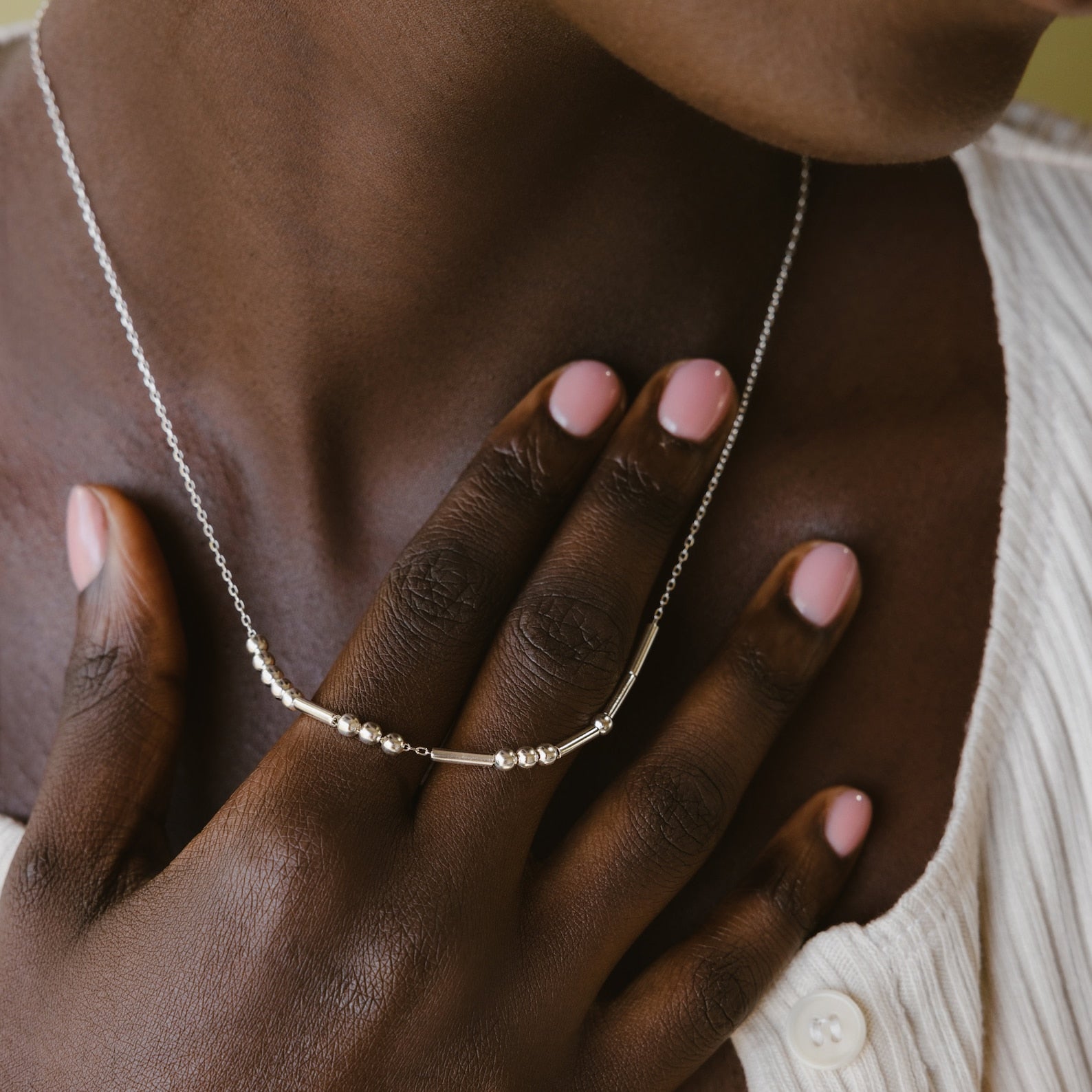 A hand with pink nails touches a Morse Code Necklace with silver beads on someone’s neck, paired with a white textured top.