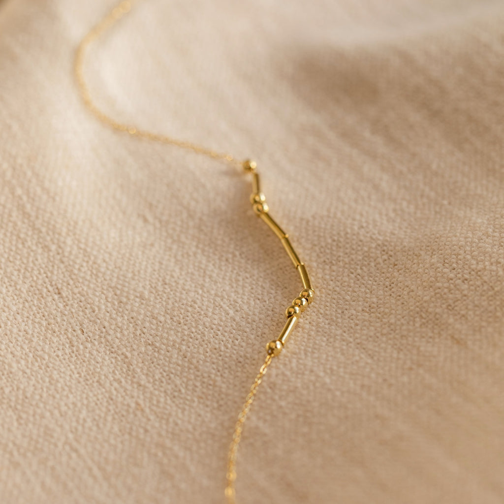 Morse Code Necklace—delicate gold beads and bars on a soft beige fabric backdrop. A perfect personalized gift.