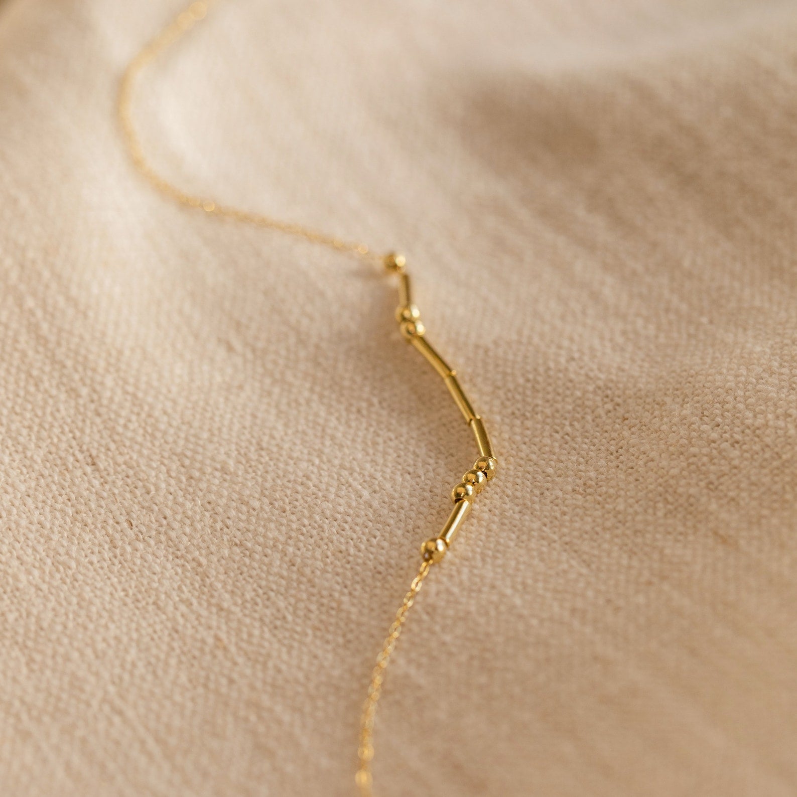 Morse Code Necklace—delicate gold beads and bars on a soft beige fabric backdrop. A perfect personalized gift.