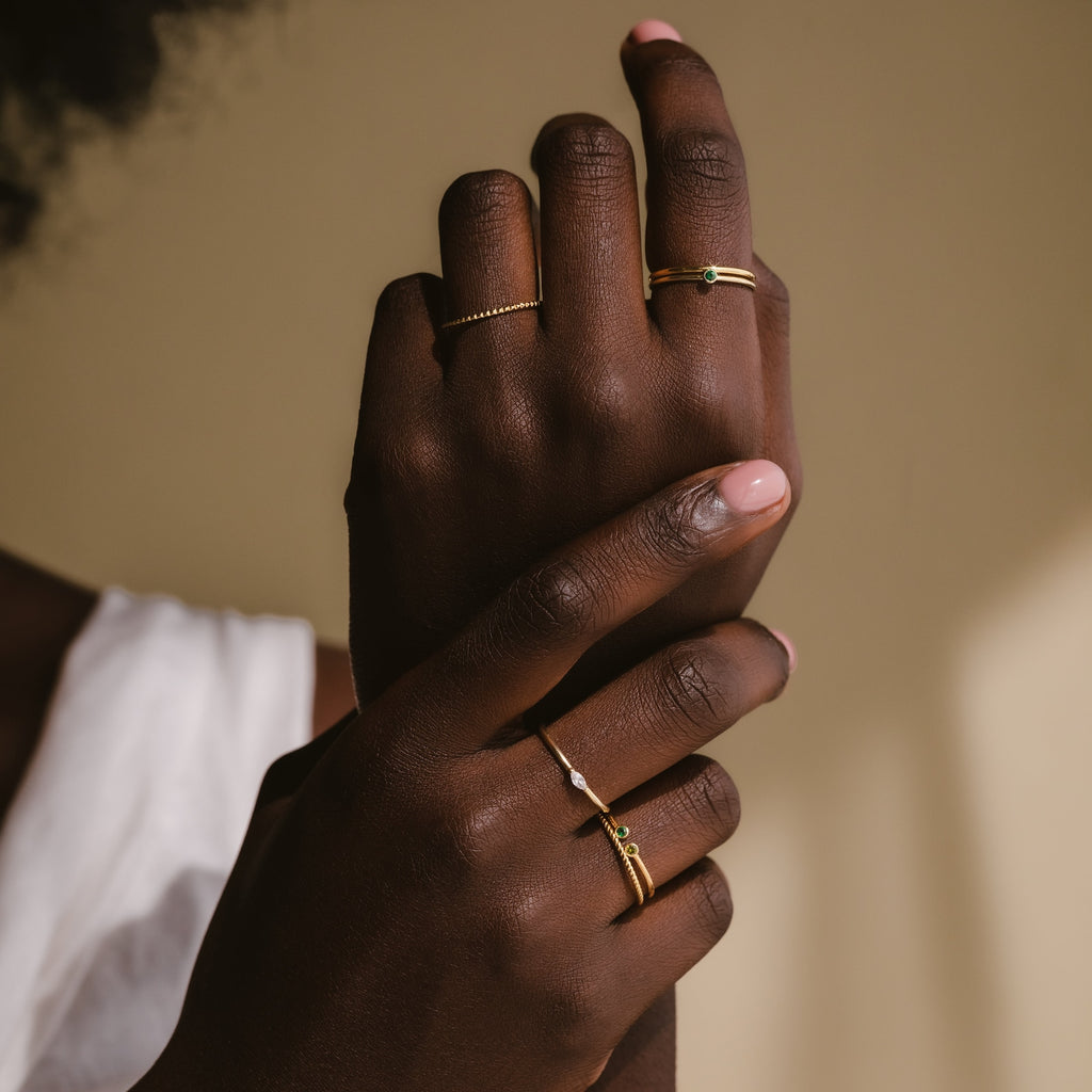 Close-up of crossed hands adorned with gold rings, featuring the Marquise Birthstone Ring, against a neutral background and styled with a white sleeveless top.