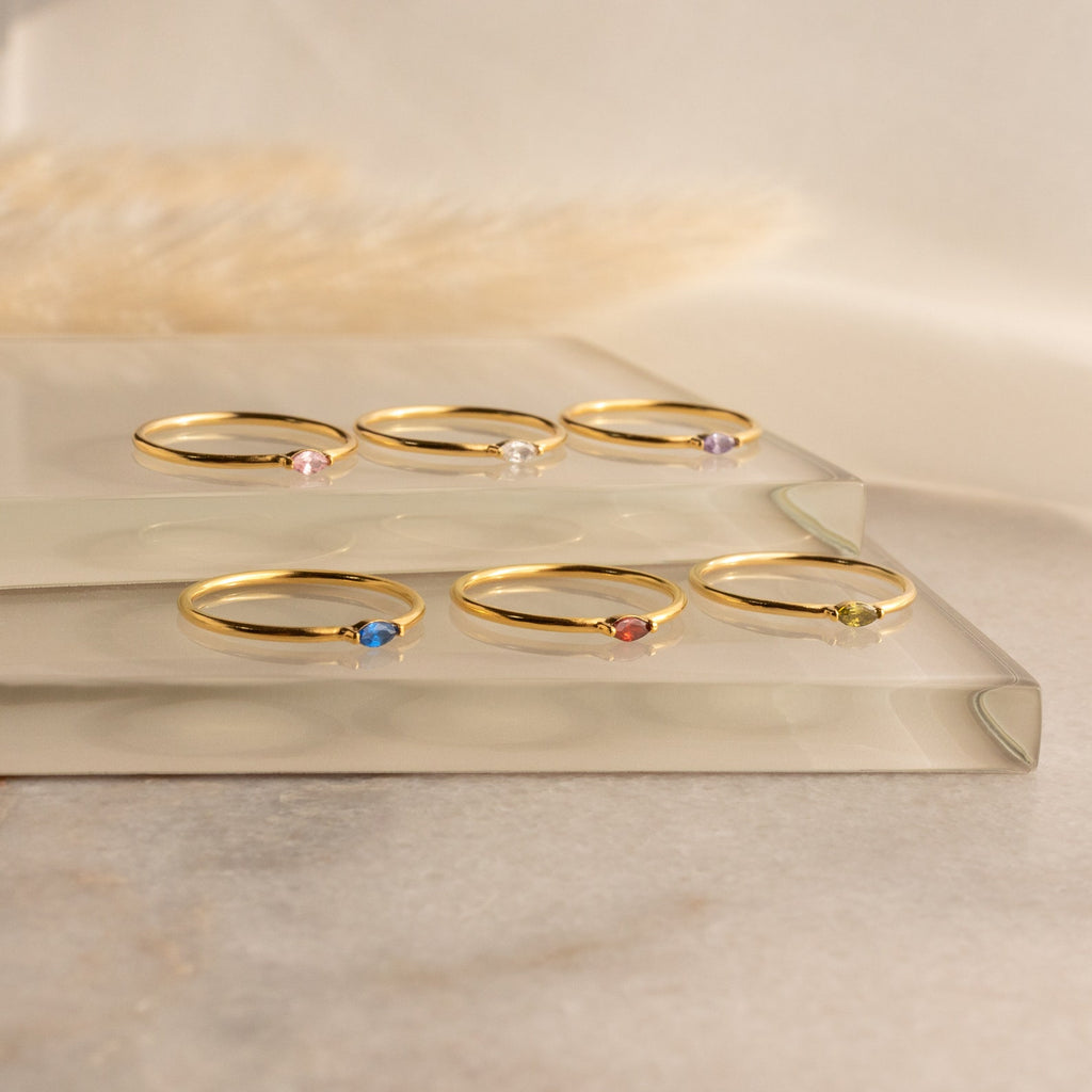 Six Marquise Birthstone Rings with colorful gemstones are showcased on two clear glass platforms against a soft background.