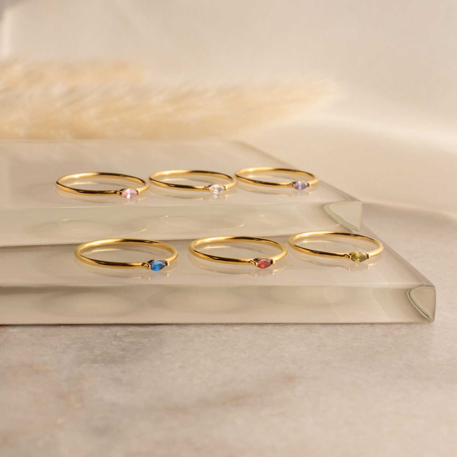 Six Marquise Birthstone Rings with colorful gemstones are showcased on two clear glass platforms against a soft background.
