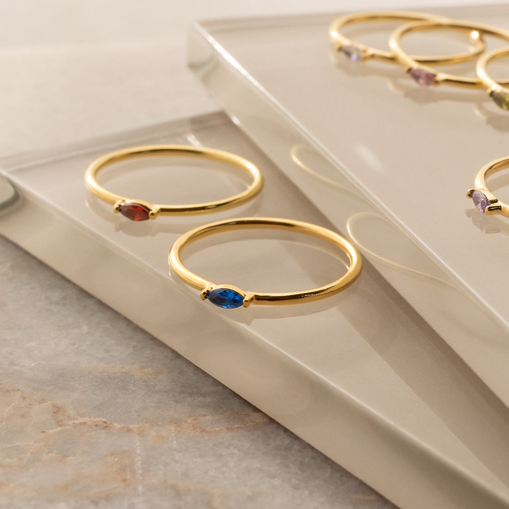 The Marquise Birthstone Ring and two gold dainty rings with colorful gemstones are displayed on reflective glass surfaces.