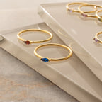 The Marquise Birthstone Ring and two gold dainty rings with colorful gemstones are displayed on reflective glass surfaces.