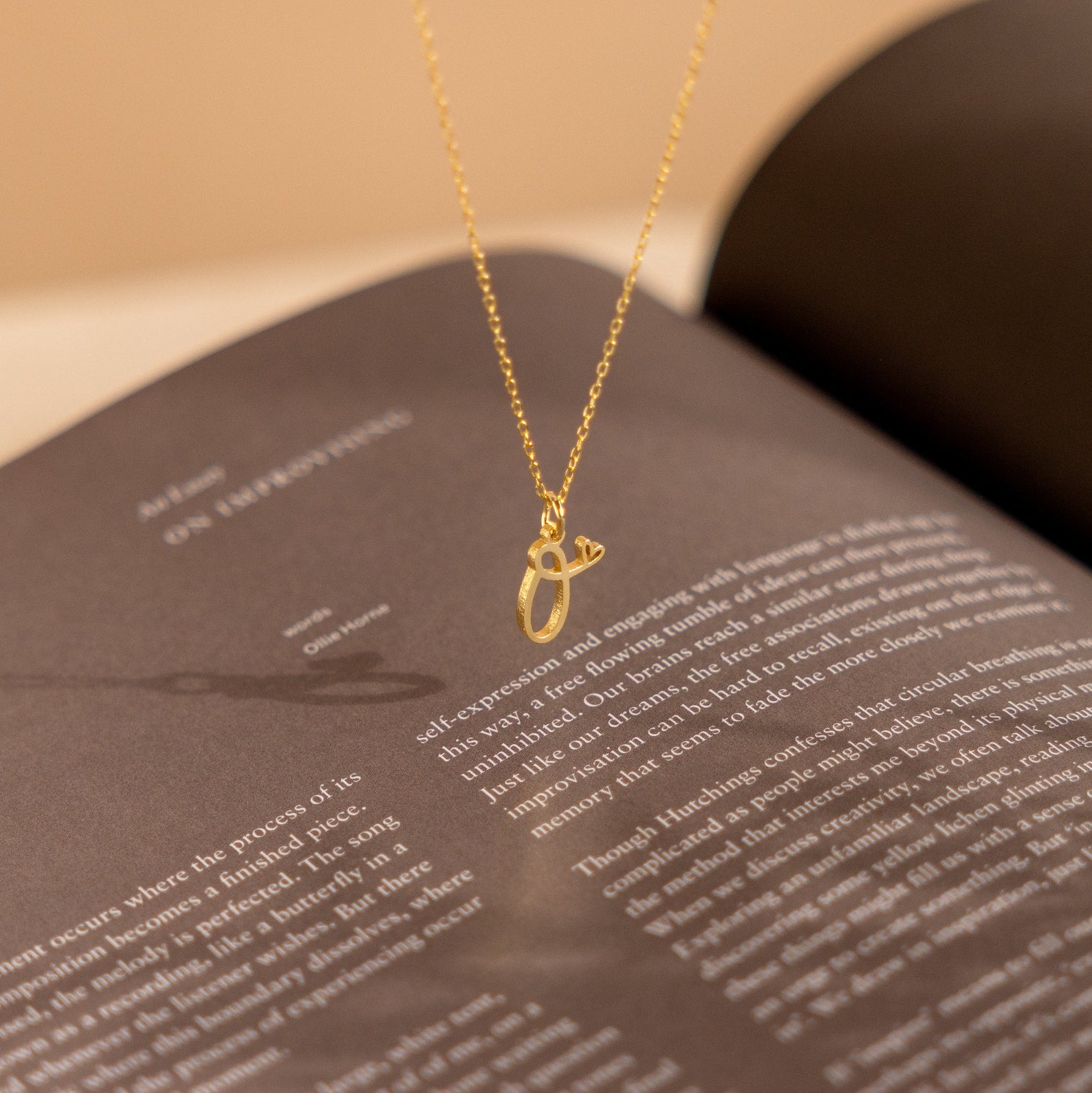 The Letter Heart Necklace, a minimalist piece featuring an abstract gold pendant, rests on an open book with blurred text.