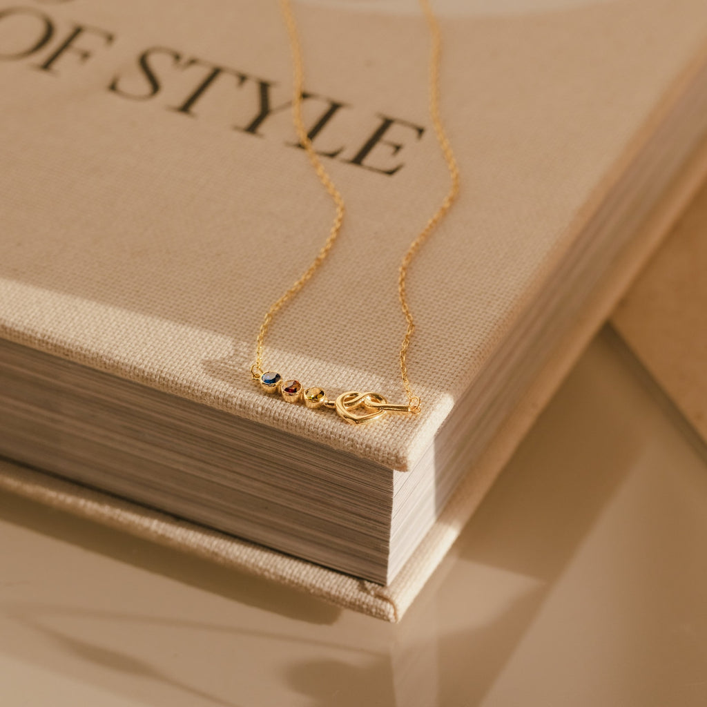 The Love Knot Birthstone Necklace with vibrant gemstone charms is elegantly displayed on the corner of a beige hardcover book.