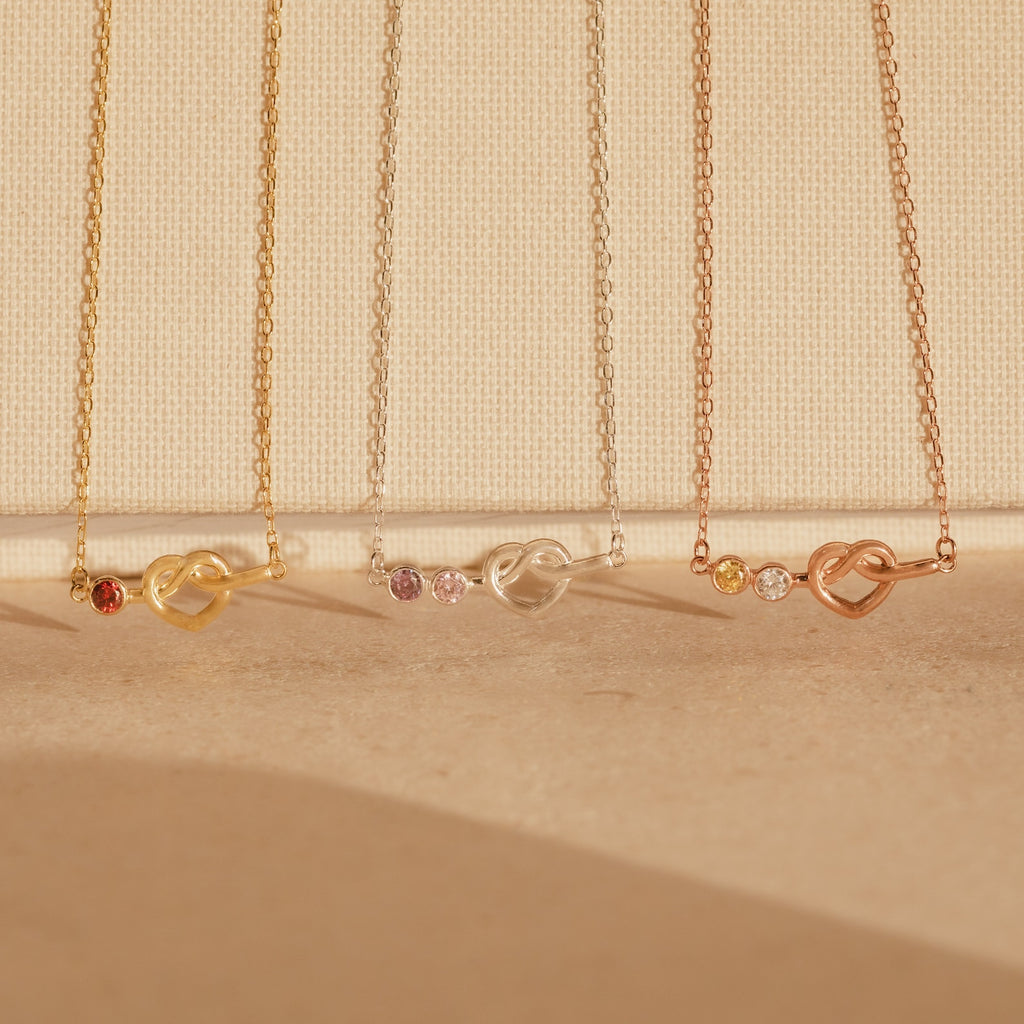 Four delicate Love Knot Birthstone Necklaces in gold, silver, and rose gold with small gemstones, perfect for celebrating family, displayed on a beige background.