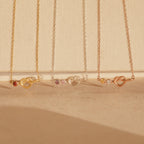 Four delicate Love Knot Birthstone Necklaces in gold, silver, and rose gold with small gemstones, perfect for celebrating family, displayed on a beige background.