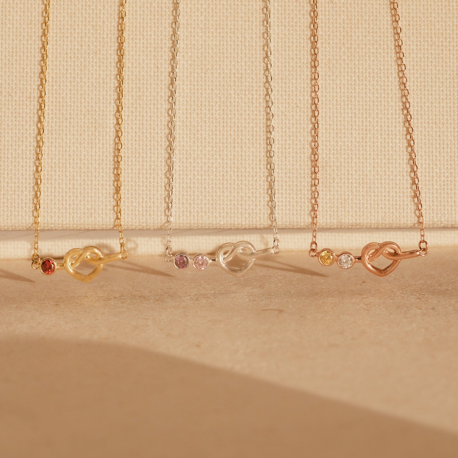 Four delicate Love Knot Birthstone Necklaces in gold, silver, and rose gold with small gemstones, perfect for celebrating family, displayed on a beige background.