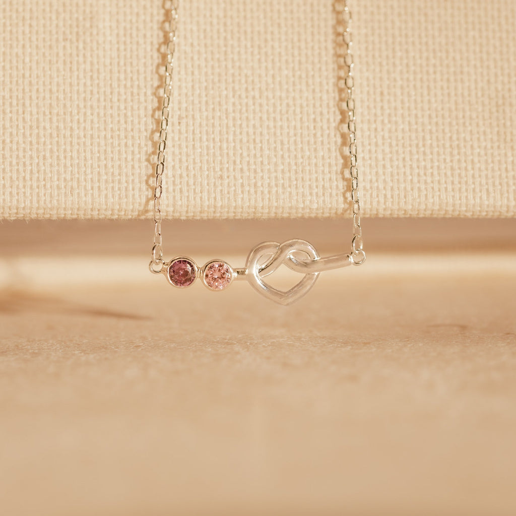 The Love Knot Birthstone Necklace features a heart-shaped pendant with two pink gems on a fine chain, set against a beige background—an elegant personalized gift to celebrate loved ones.