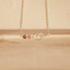 The Love Knot Birthstone Necklace features a heart-shaped pendant with two pink gems on a fine chain, set against a beige background—an elegant personalized gift to celebrate loved ones.