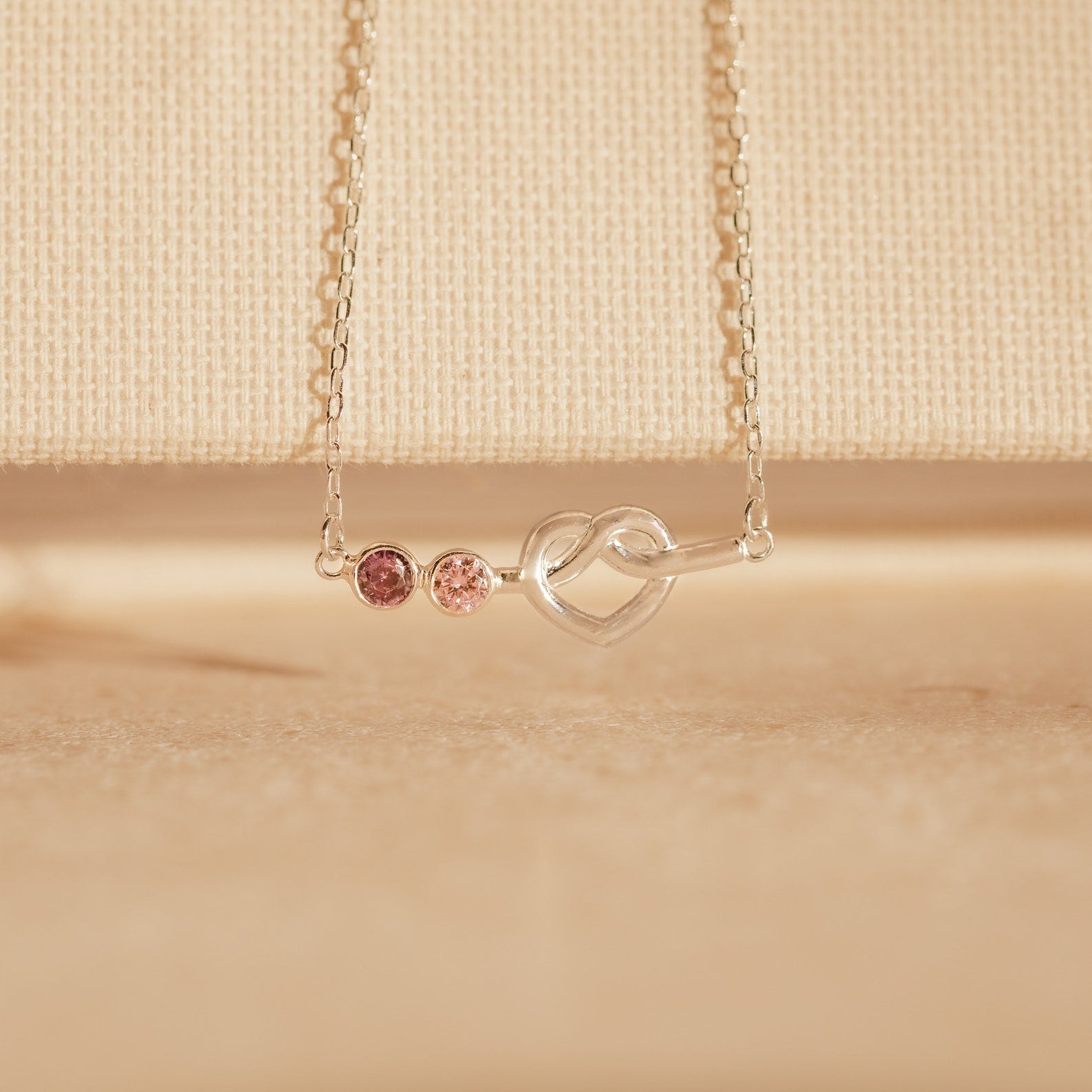 The Love Knot Birthstone Necklace features a heart-shaped pendant with two pink gems on a fine chain, set against a beige background—an elegant personalized gift to celebrate loved ones.