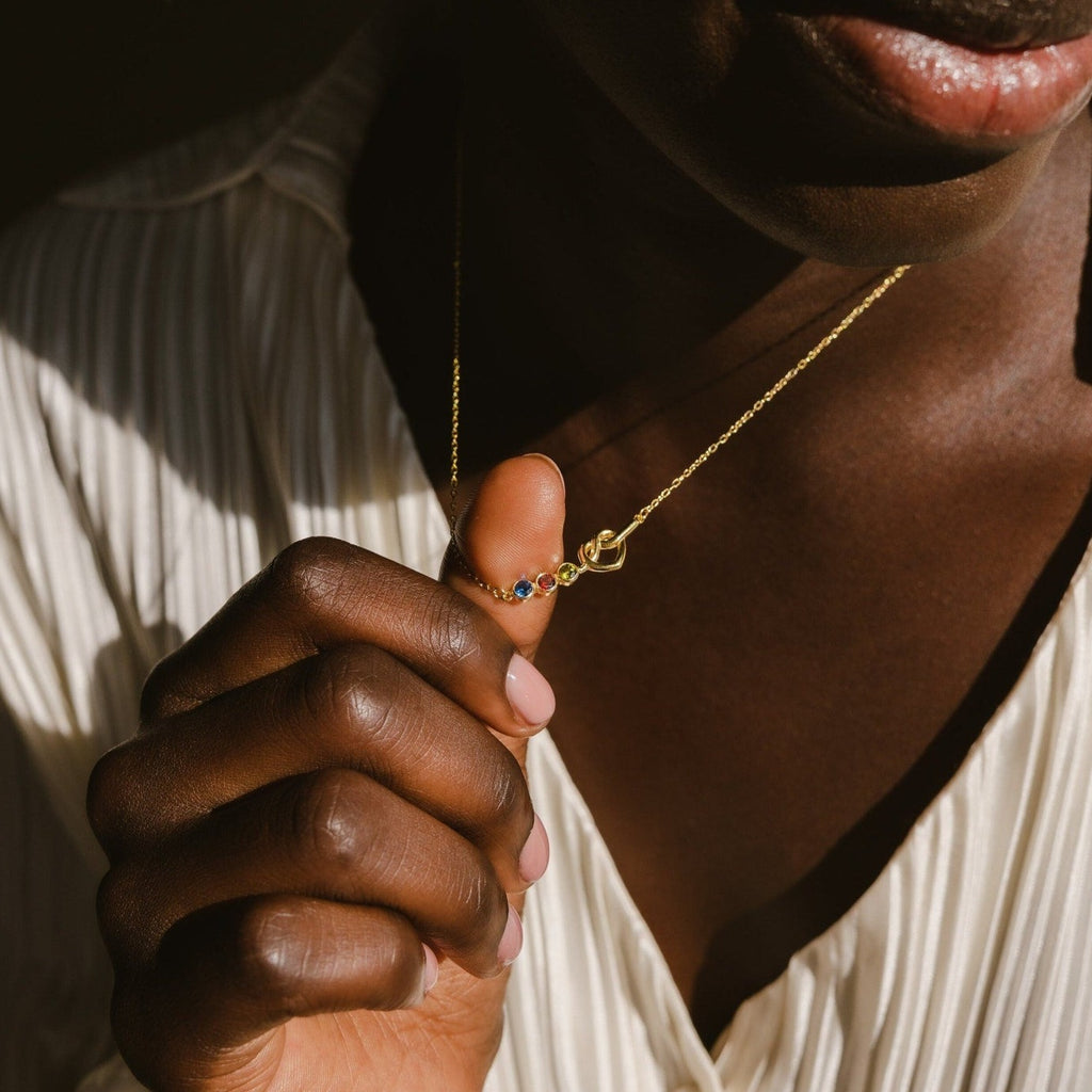 A person wearing a cream pleated top holds the Love Knot Birthstone Necklace, a delicate gold piece adorned with vibrant gemstones.