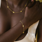 A woman, dressed in a cream-colored top, wears a Flower Station Necklace and matching bracelet featuring gold floral designs.