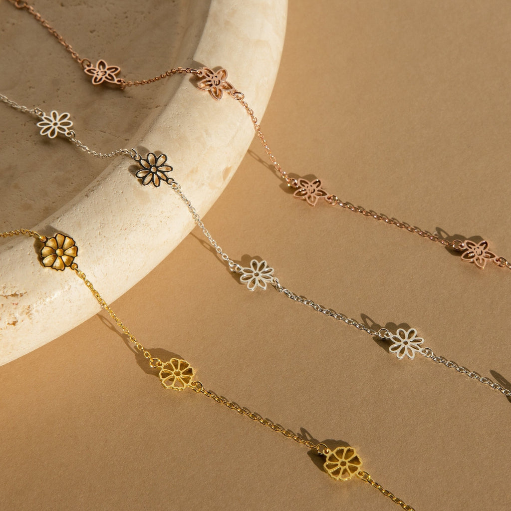 Our Flower Station Necklace, a delicate floral chain, is shown in gold, silver, and rose gold on a beige stone surface—a beautiful piece of jewelry and an elegant gift for her.
