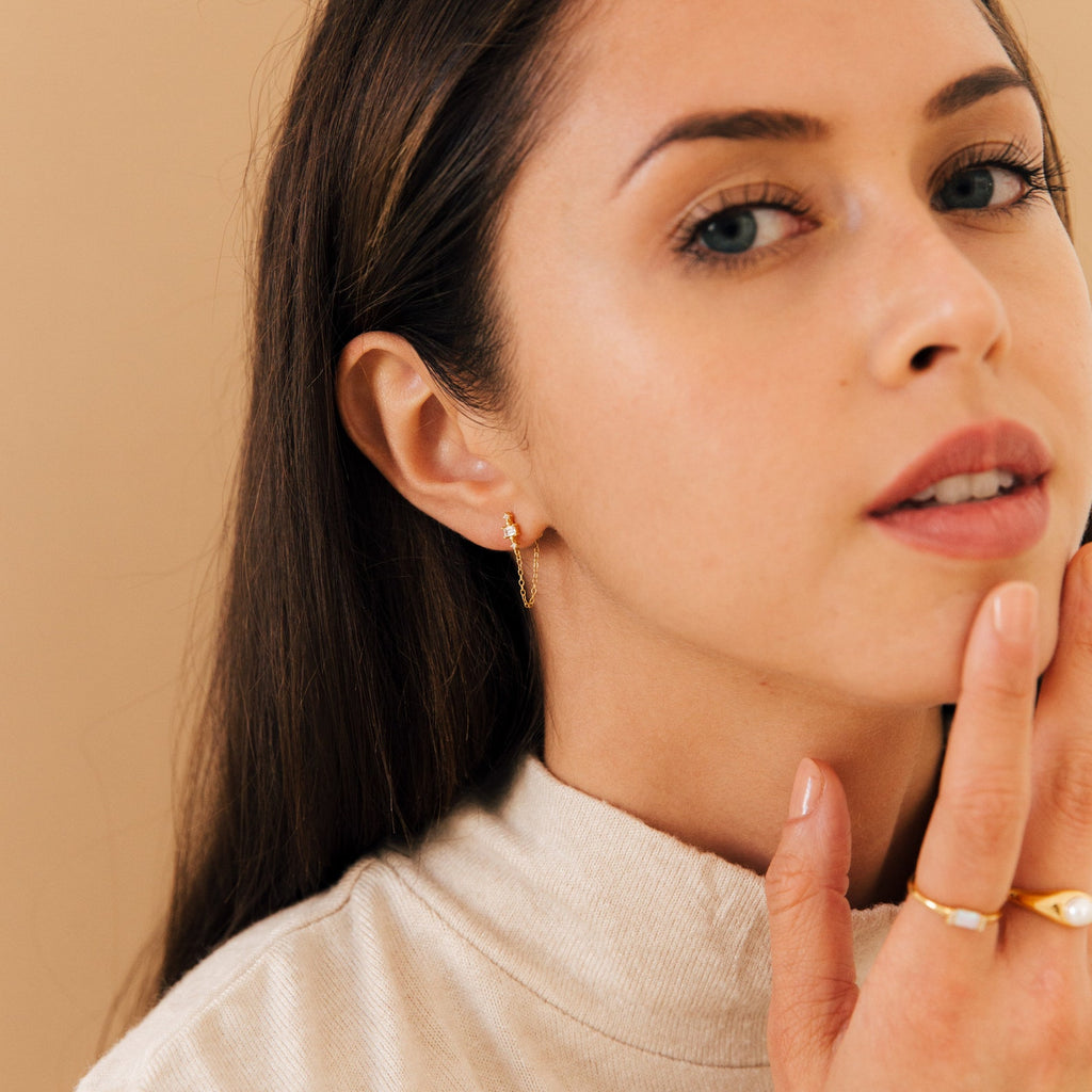 Woman with long brown hair wears Florence Diamond Chain Earrings and a ring, touches her face, and looks at the camera.