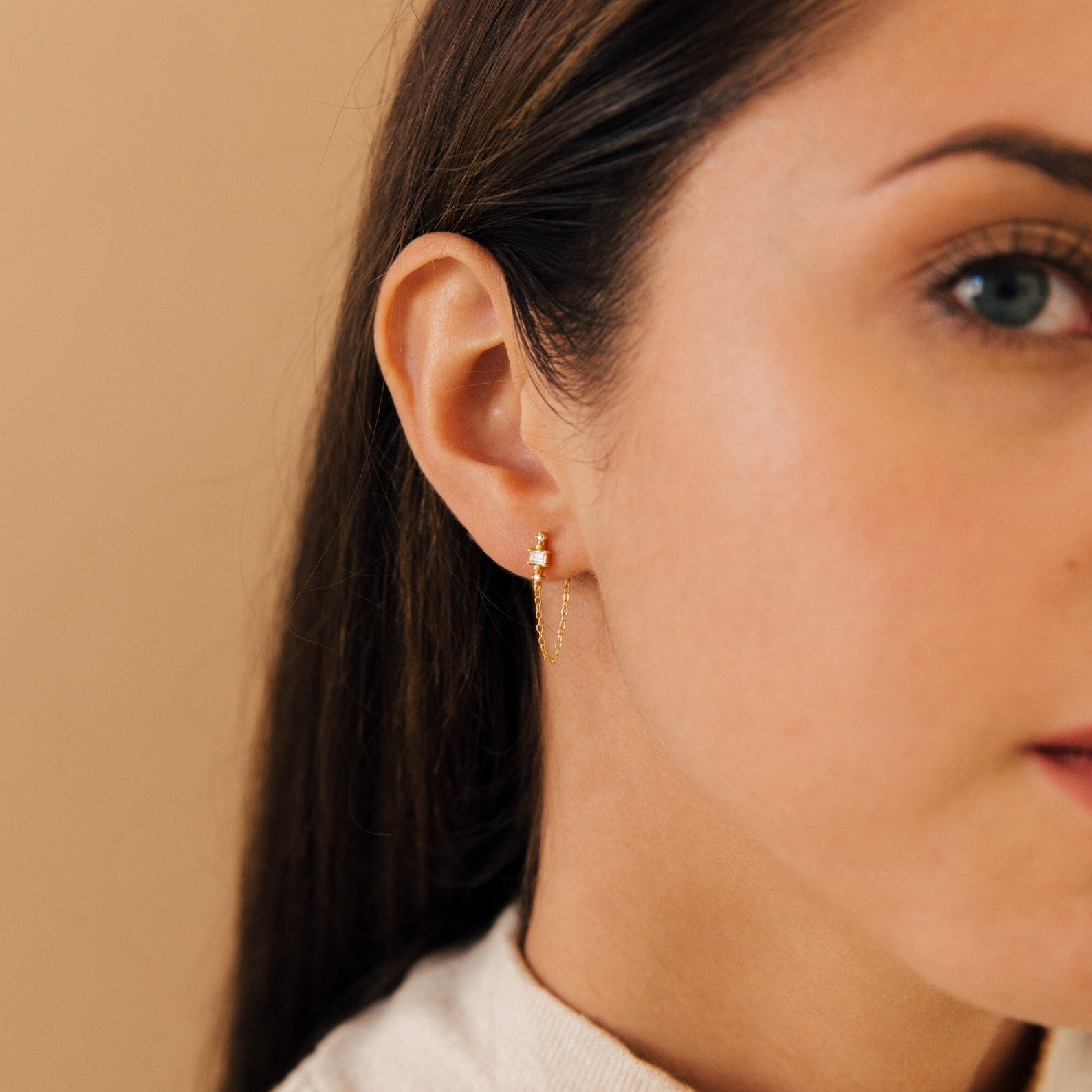 A woman with straight brown hair wears Florence Diamond Chain Earrings, shown in close-up against a beige background.