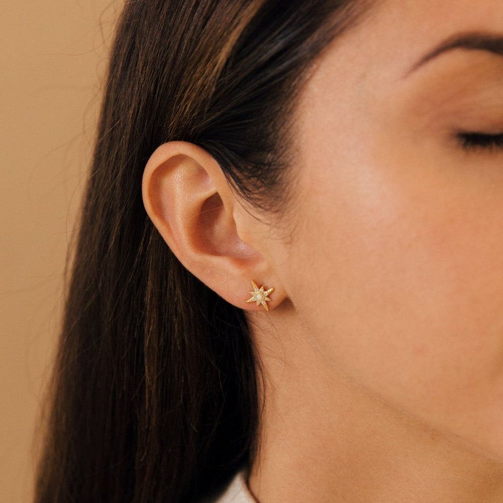 Close-up of a woman’s ear wearing the Starburst Studs—small gold earrings with a clear gemstone center.