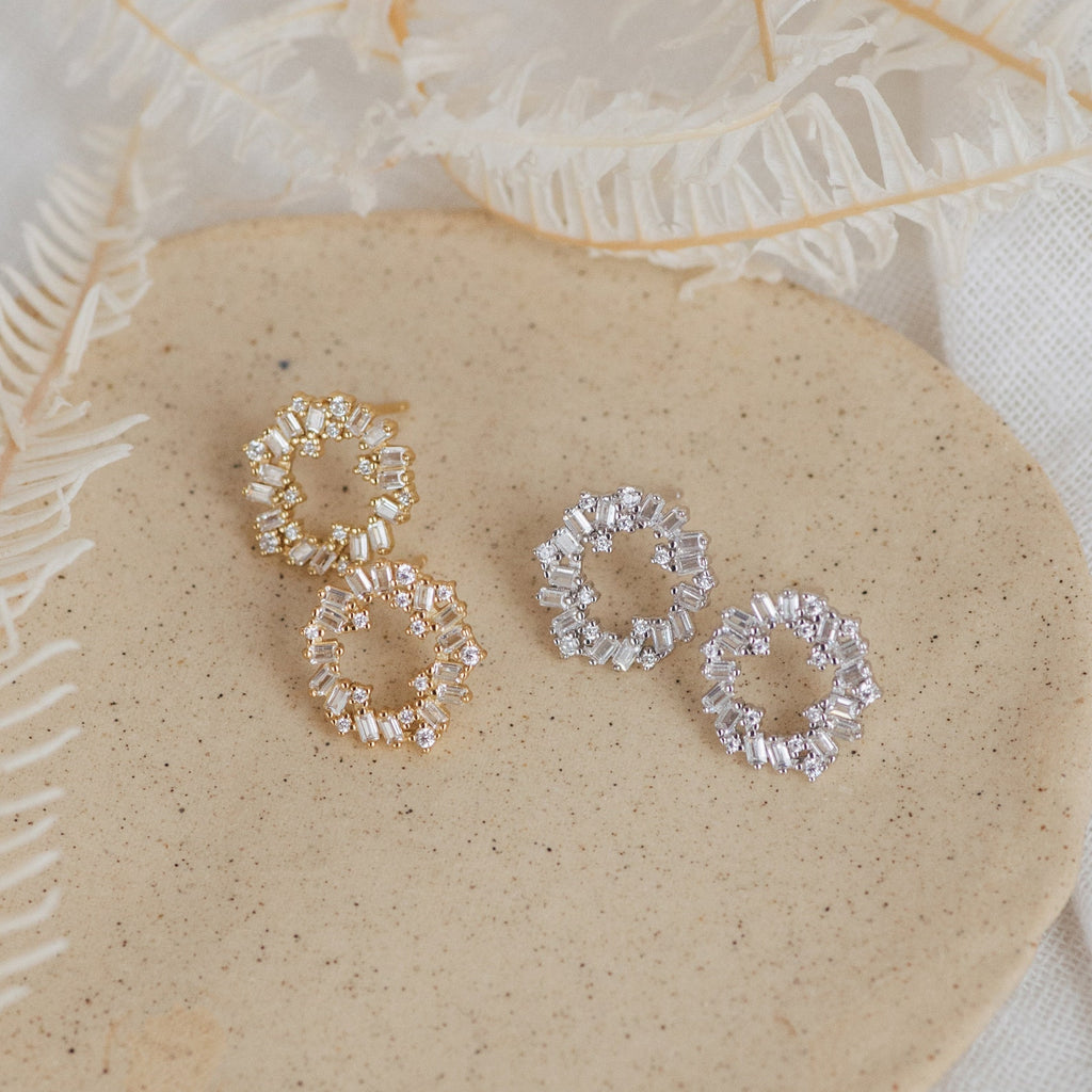 Two pairs of Reina Cluster Studs, elegant bridal earrings in gold and silver, are displayed on a beige surface with dried foliage.