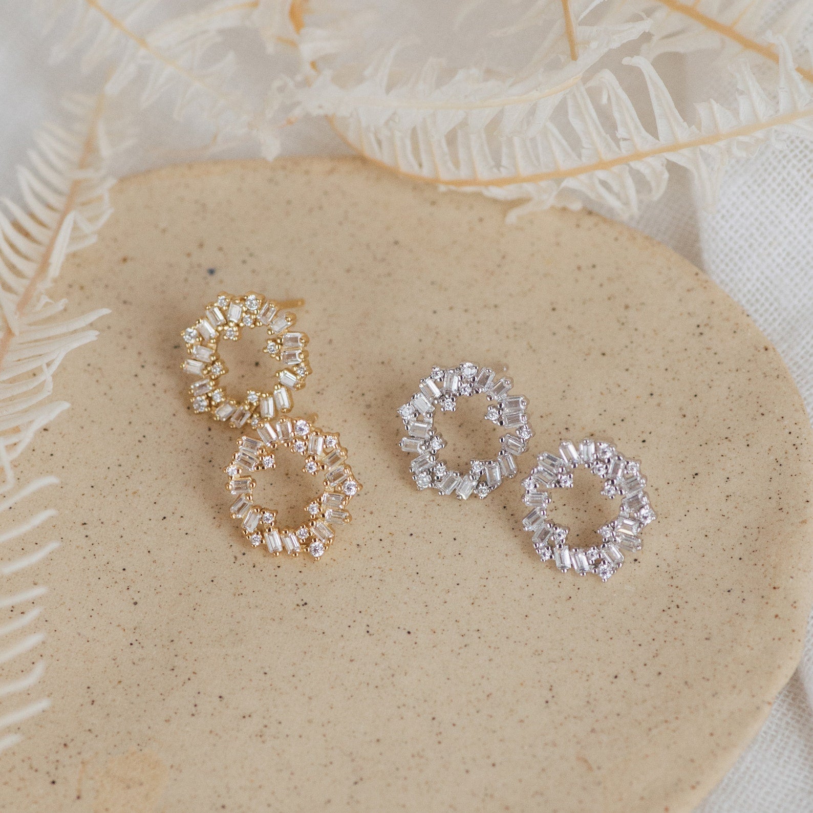 Two pairs of Reina Cluster Studs, elegant bridal earrings in gold and silver, are displayed on a beige surface with dried foliage.