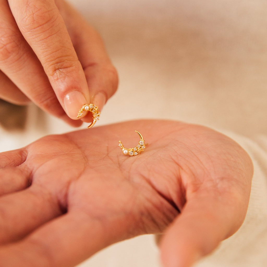 A hand holds a small gold hoop diamond pearl earring, while another hand picks up the matching Eclipse Moon Stud earring.
